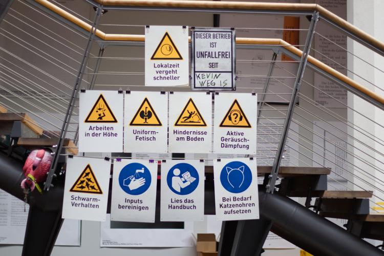 Several warning signs, featuring texts such as "Lokalzeit vergeht schneller", "Aktive Ger&auml;uschd&auml;mpfung", "Schwarmverhalten" and "Bei Bedarf Katzenohren aufsetzen"